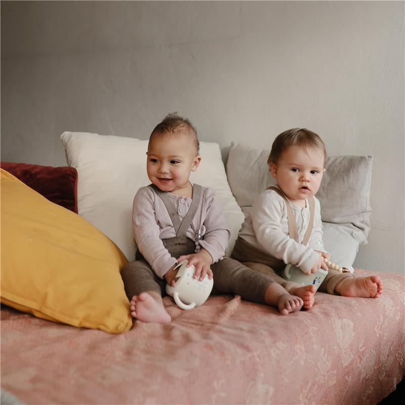 Two babies sit on a couch, one holding a white Mushie snack cup while wearing a light purple outfit, and the other holds a blue snack cup, both smiling.