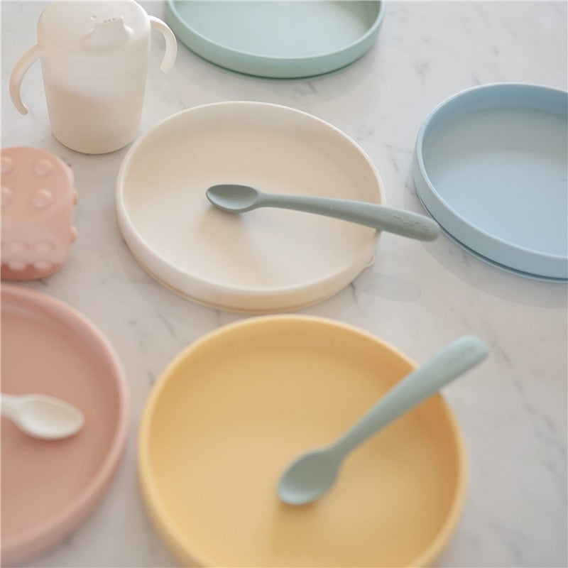 Mushie Classic Silicone Suction Plate in Powder Blue with a light green spoon, alongside other colorful silicone plates and a cup.