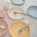 Mushie Classic Silicone Suction Plate in Powder Blue with a light green spoon, alongside other colorful silicone plates and a cup.