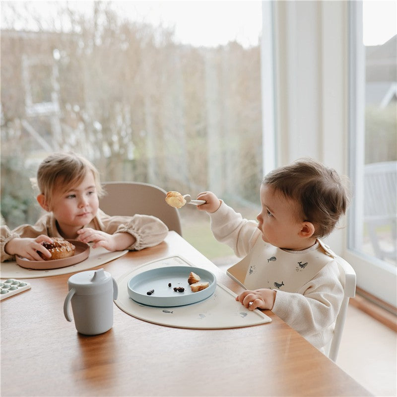 Mushie Classic Silicone Suction Plate in Powder Blue, with a child using a spoon to eat while seated at a dining table.