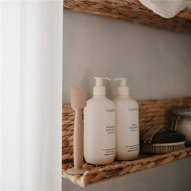Mushie silicone diaper cream applicator next to shampoo and body wash bottles on a woven shelf.