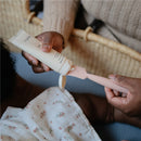 Mushie silicone diaper cream applicator held by a person, dispensing cream onto the applicator while a baby in a floral cloth is nearby.