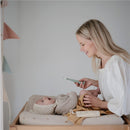 A mother smiles while using the Mushie silicone diaper cream applicator on her baby, who is on a changing table.
