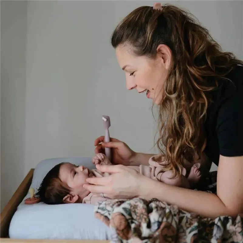 Mother uses a soft lilac Mushie diaper cream applicator with suction base while interacting with her baby on a changing table.