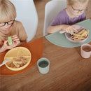 Two children eat from colorful silicone plates and cups at a wooden table, showcasing the Mushie Silicone Dinnerware Cup Set.