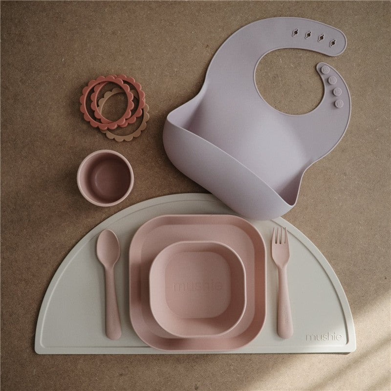 Mushie silicone dinnerware set in blush, featuring a fork, spoon, plates, a cup, a bib, and stackable rings.
