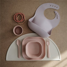 Mushie silicone dinnerware set in blush, featuring a fork, spoon, plates, a cup, a bib, and stackable rings.
