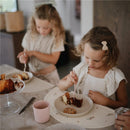 Two children enjoying dessert using a pink Mushie silicone fork and spoon set on a table with cake and cookies.