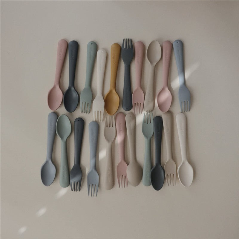 Mushie silicone dinnerware fork and spoon set in various pastel colors displayed on a neutral background.