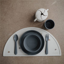 Mushie Silicone Dinnerware Fork & Spoon Set in Smoke, featuring a fork, spoon, bowl, plate, and cup on a placemat.