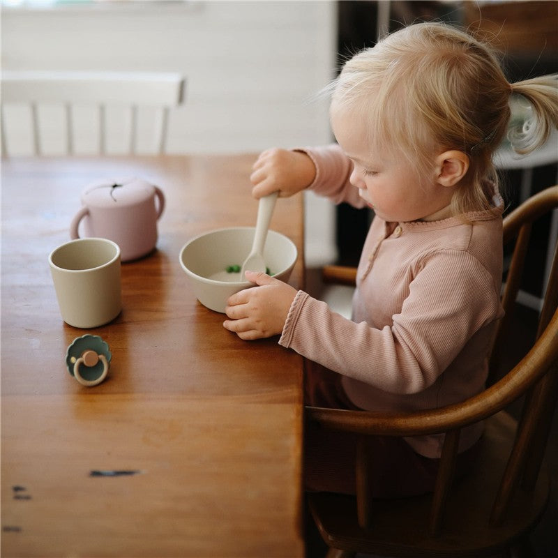 A toddler with blonde hair uses a silicone spoon to stir food in a bowl at a wooden table, with a cup and teapot nearby.