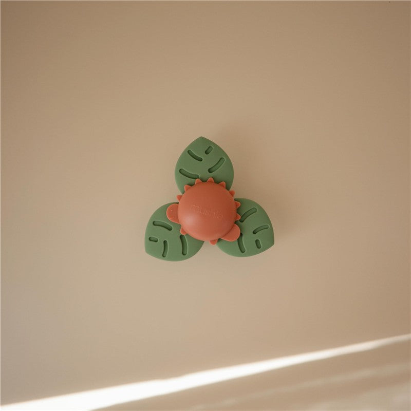 Mushie Dino Suction Spinner Toy featuring a central orange dinosaur surrounded by green leaf-shaped pieces for bath fun.