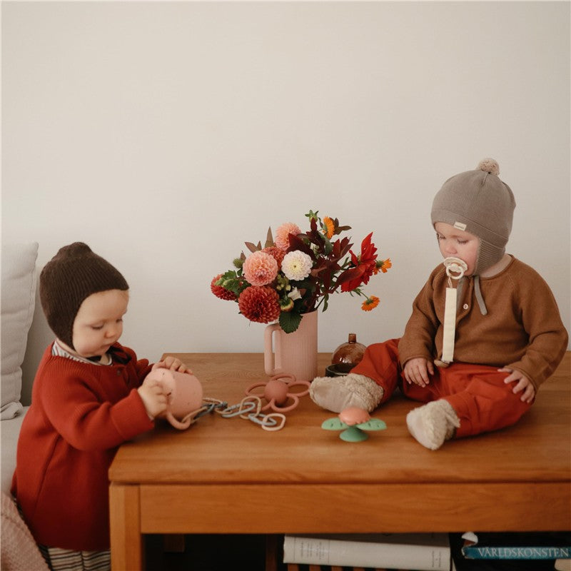 Two toddlers play on a wooden table, one interacting with a suction spinner toy, while a flower vase decorates the scene.