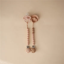 Mushie Eva Pacifier Clip in blush, featuring two clips with beads, a silicone pacifier with a floral design, and a wooden clip.