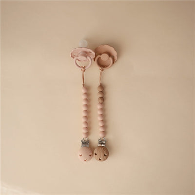 Mushie Eva Pacifier Clip in blush, featuring two clips with beads, a silicone pacifier with a floral design, and a wooden clip.