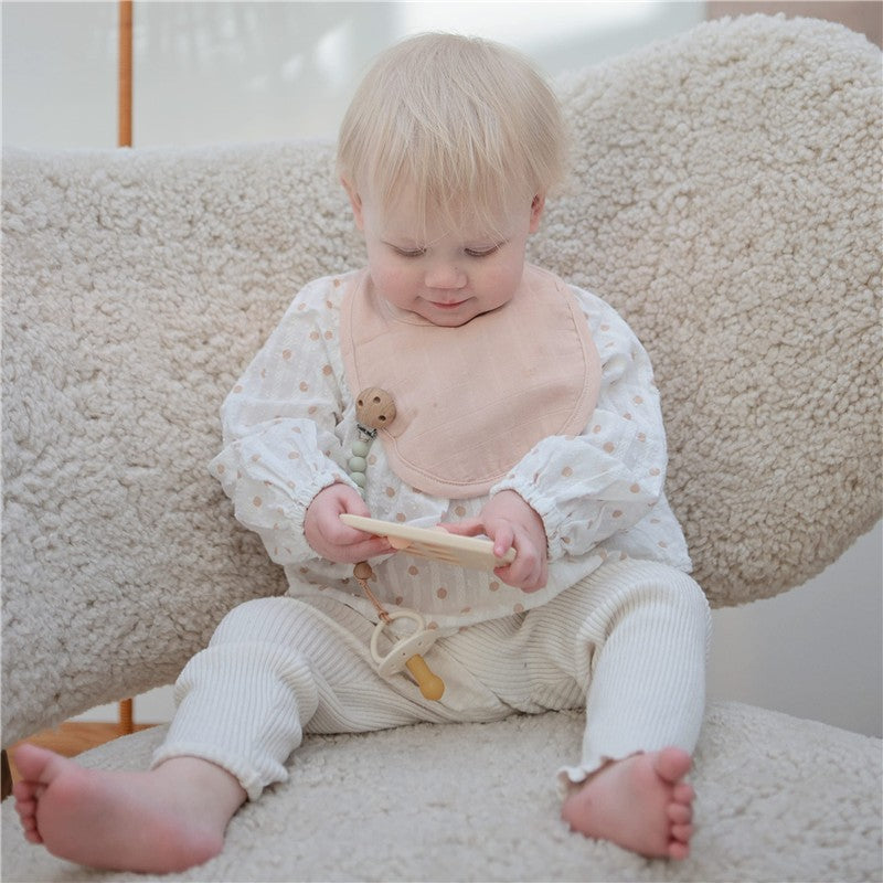 A happy baby wearing a polka dot blouse and pink bib sits in a cozy chair, holding a wooden toy with a Mushie Eva Pacifier Clip attached.