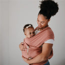 Mother gently holds a baby wrapped in a soft fabric carrier, both smiling, with the baby wearing a headband.