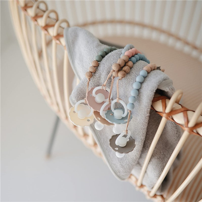 Mushie Eva Pacifier Clips in cloud colors hang from a gray towel draped over a wooden crib, featuring pastel beads and cloud-shaped clips.