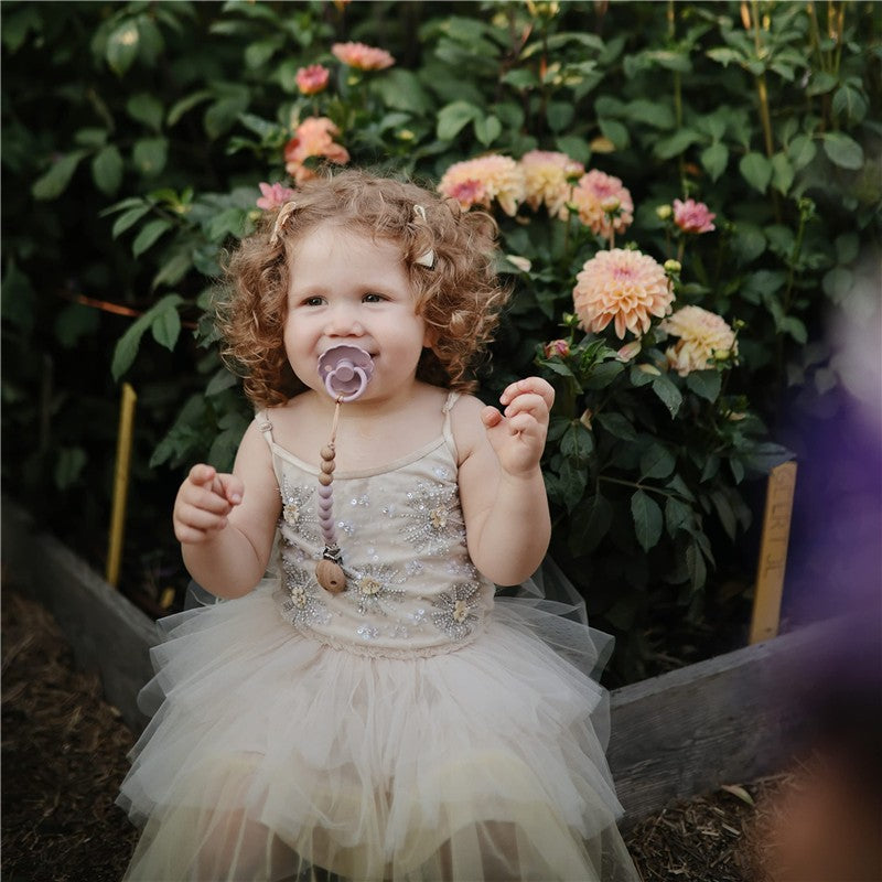 Toddler wearing a sparkly dress with a mauve pacifier clip, sitting amidst colorful flowers, smiling and playing.