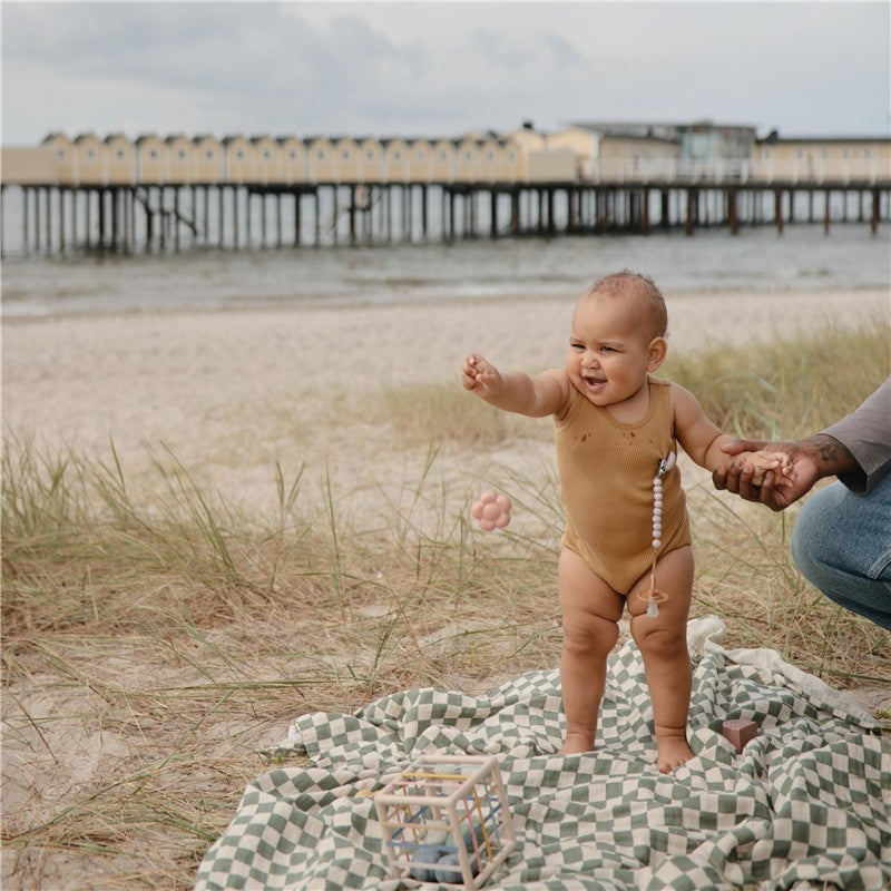 A smiling baby wearing a brown onesie stands on a grassy beach blanket, holding a hand and reaching for a toy, with a pier in the background.