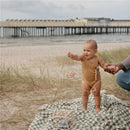 A smiling baby wearing a brown onesie stands on a grassy beach blanket, holding a hand and reaching for a toy, with a pier in the background.