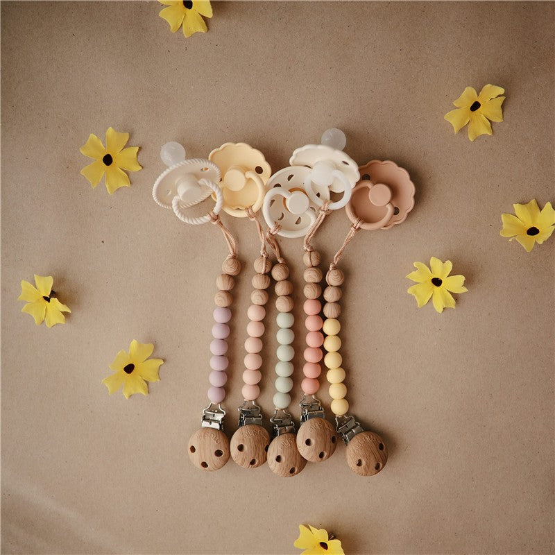 Mushie Eva Pacifier Clip in mauve with wooden beads and a floral pacifier holder, surrounded by yellow paper flowers.