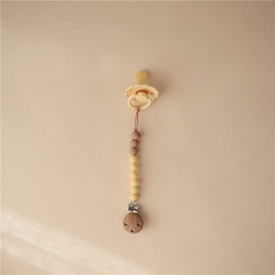 Muted yellow Mushie Eva pacifier clip with wooden beads and a clip, designed to secure pacifiers.
