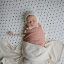 Ivory Mushie Extra Soft Baby Blanket drapes softly around a sleeping baby, who wears a beige swaddle and a knitted cap.