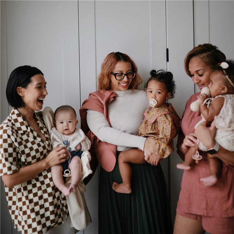 Four women smile while holding babies; the focus is on a baby blanket. The product is Mushie Extra Soft Baby Blanket in Ivory.