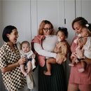 Four women smile while holding babies; the focus is on a baby blanket. The product is Mushie Extra Soft Baby Blanket in Ivory.