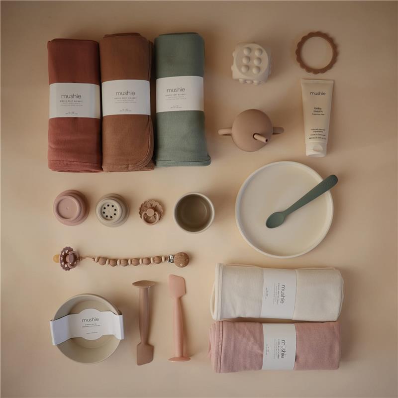Mushie Extra Soft Baby Blanket in ivory, alongside additional baby items like bowls, spoons, and toys in soft colors.