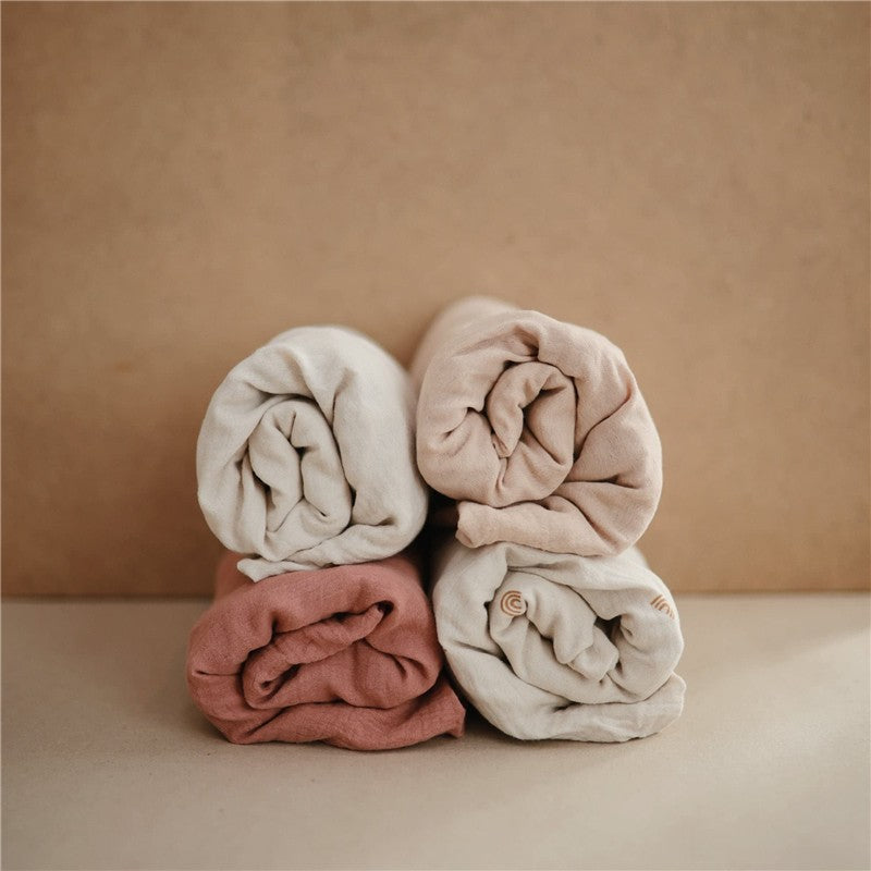 Four rolled muslin changing pad covers in soft colors: cream, light pink, and terracotta, stacked neatly on a neutral background.