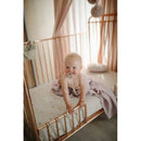 A toddler with a pacifier sits on a mini crib sheet in a rose gold crib, surrounded by soft pink decor.