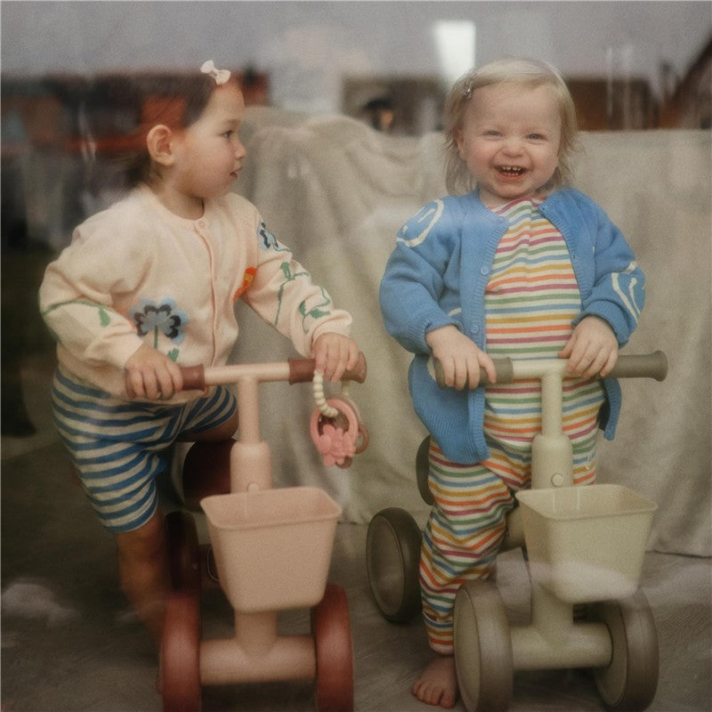 Two toddlers play with toy ride-on tricycles in a cozy setting, one holding a teething ring.