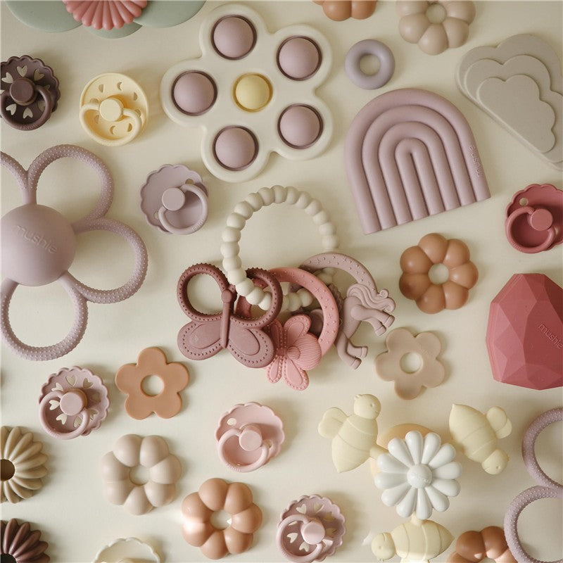 Mushie Fairy Teething Ring surrounded by various colorful silicone toys and teething accessories in soft pastel shades.