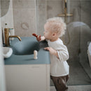 Child using a Mushie 2-Pack Finger Toothbrush in clay and shifting sand colors, leaning against a bathroom sink.