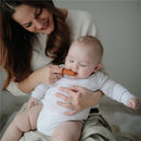 Baby using a clay-colored Mushie finger toothbrush while being held by a smiling caregiver, both sitting on a light-colored surface.