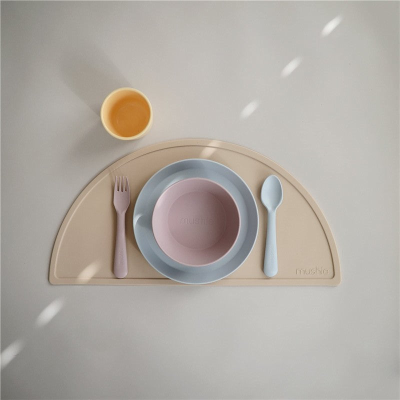 Mushie silicone fork and spoon set in powder blue with a pink bowl and yellow cup on a beige placemat.