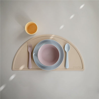 Mushie silicone fork and spoon set in powder blue with a pink bowl and yellow cup on a beige placemat.