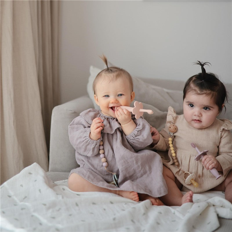Two babies sitting on a sofa, one happily mouthing a pink silicone toothbrush while the other holds a similar toothbrush.