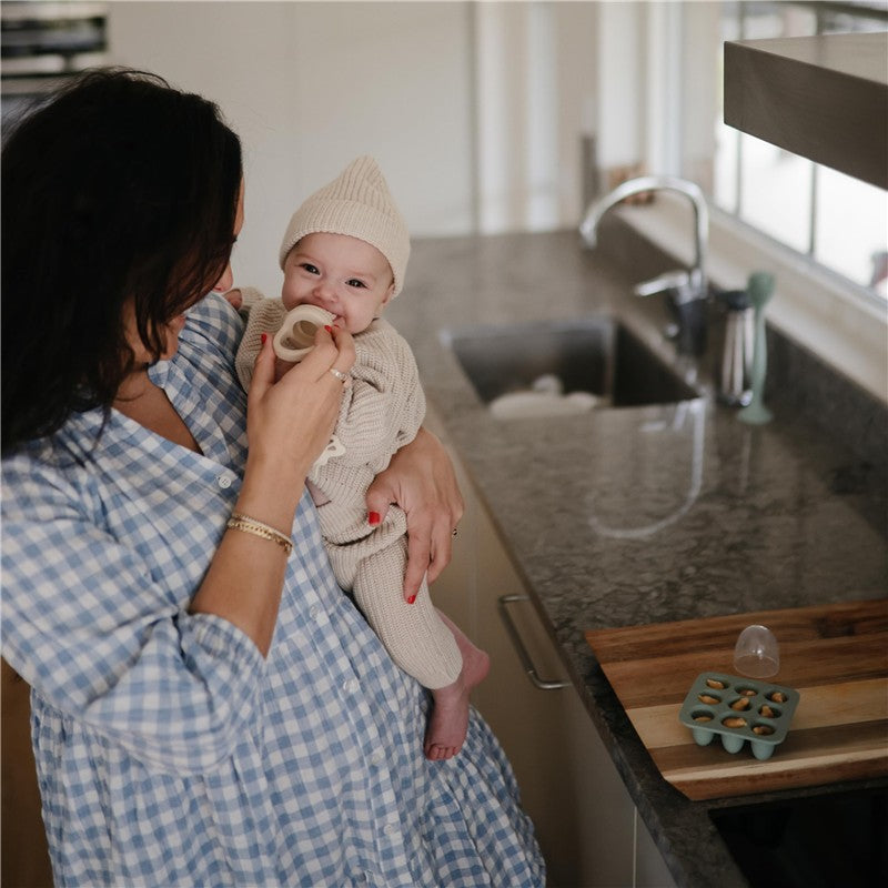 A woman in a blue checkered dress holds a smiling baby wearing a knitted outfit and hat, near a granite kitchen counter with a Mushie Fresh Food Feeder Freezer Tray.