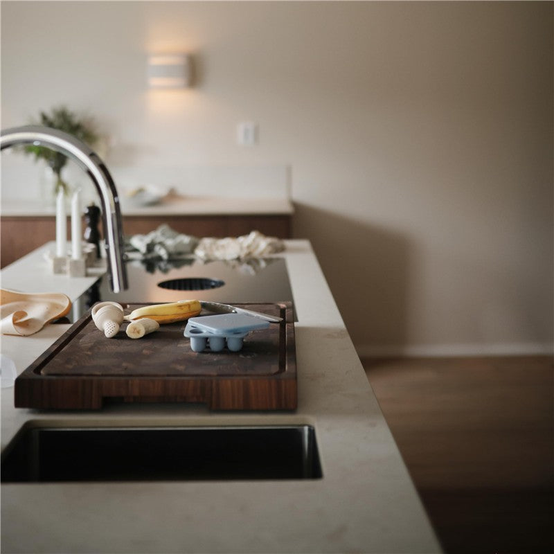 Mushie Fresh Food Feeder Freezer Tray in light blue on a wooden cutting board, with fruits and a knife in a modern kitchen.
