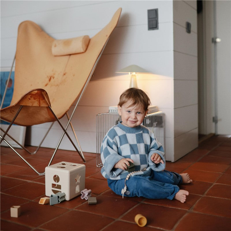 Child sitting on a tiled floor, wearing a blue and white sweater, playing with geometric blocks and a silicone pacifier clip.