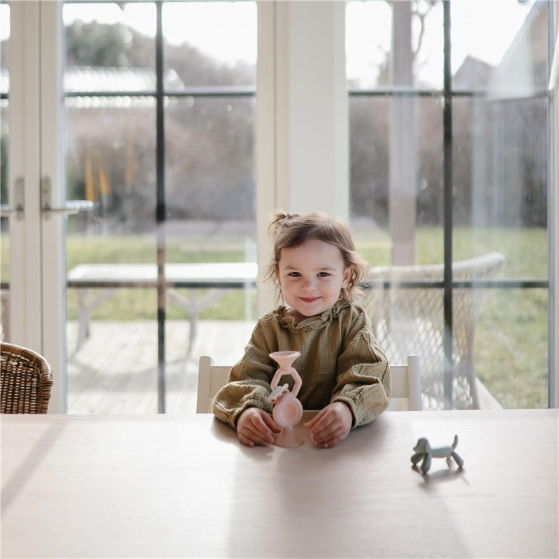 A smiling child holds a blush-colored Mushie Silicone Hexapop suction toy on a wooden table, with a sunny background.