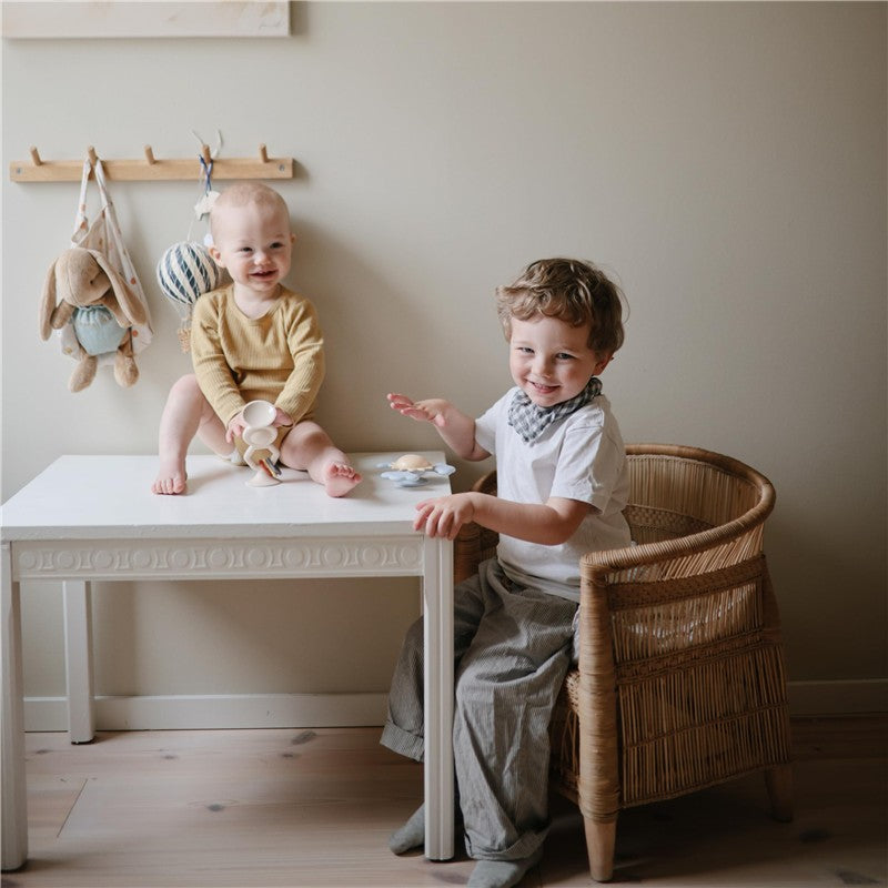 Two young children play at a small white table; one child sits on the table with a toy, while the other gestures happily.