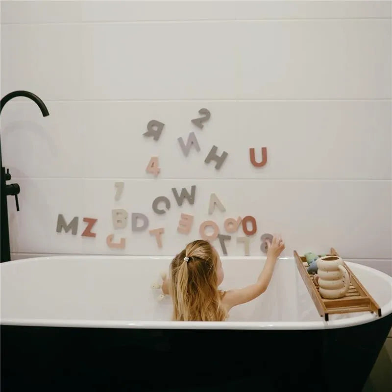 A child interacts with colorful foam letters and numbers on a bathroom wall while playing in a bathtub.