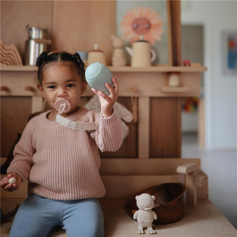 A child holds a green silicone toy, wearing a blush pacifier secured with a linen clip, seated by wooden decor.