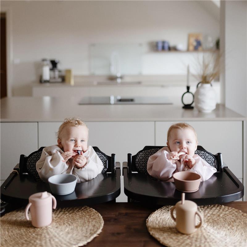Two babies in high chairs are enjoying snacks, wearing long-sleeve bibs. A gray bowl and a pink cup are on the table.