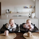 Two babies in high chairs are enjoying snacks, wearing long-sleeve bibs. A gray bowl and a pink cup are on the table.