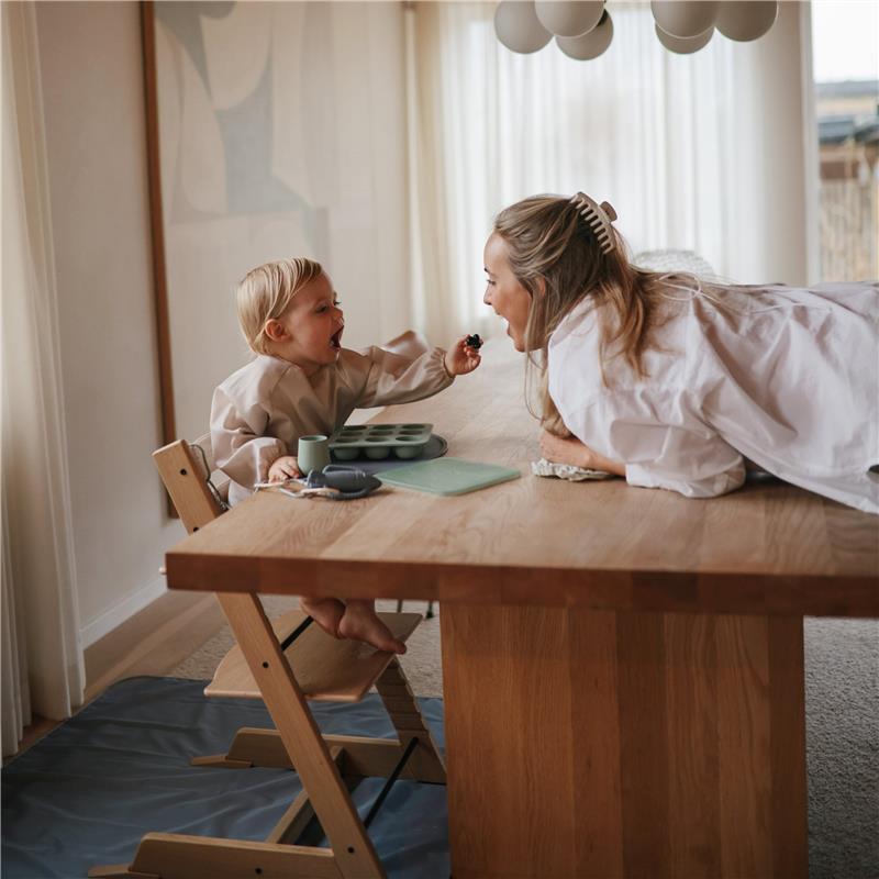A joyful toddler in a beige long sleeve bib shares food with a smiling woman at a wooden table, surrounded by dining accessories.